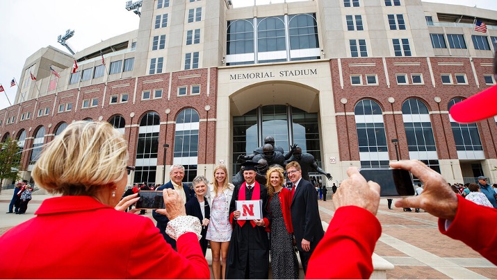 Student and his family pose in front of the Memorial Stadium during a graduation celebration. Two people are taking their photos. 