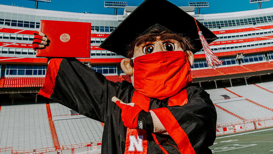 Herbie Husker wearing a mask in a graduation cap and gown pointing to a diploma