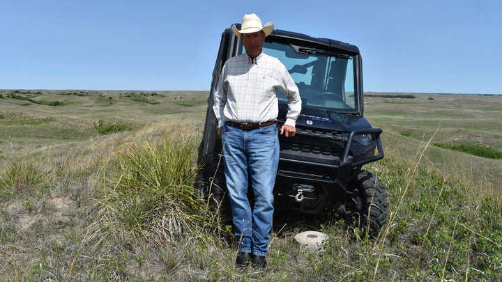 Homer Buell, owner and operator of Shovel Dot Ranch near Rose, Nebraska, stands in front of an all-terrain vehicle in a field. He is wearing a light-colored longsleeve plaid shirt, blue jeans and a white cowboy hat.