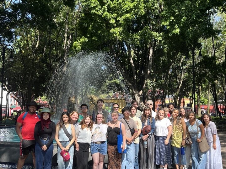 Group of people posing in front of a fountain surrounded by trees in a sunny park.